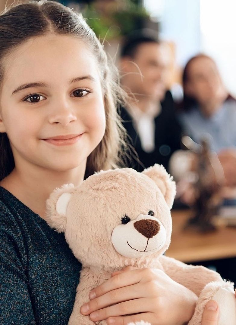 Ein kleines Mädchen mit einem Teddybär, im Hintergrund sitzen die Eltern beim Anwalt